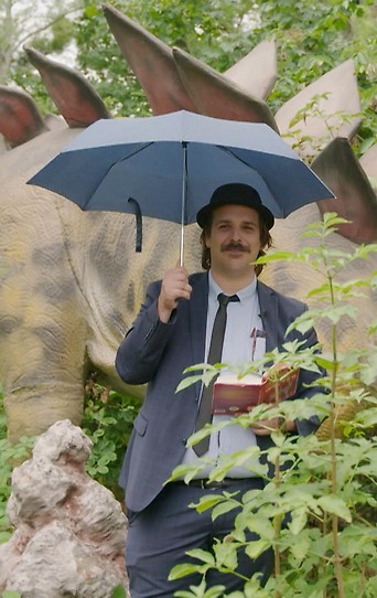 Schriftsteller Ferdinand Schmalz mit Regenschirm vor einem Dinosaurier im Wald