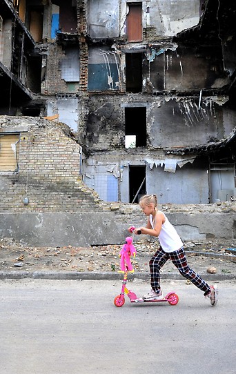 Junges Mädchen auf einem Roller vor einer zerstörten Häuserfront im Dorf Horenka in der Region Kiew