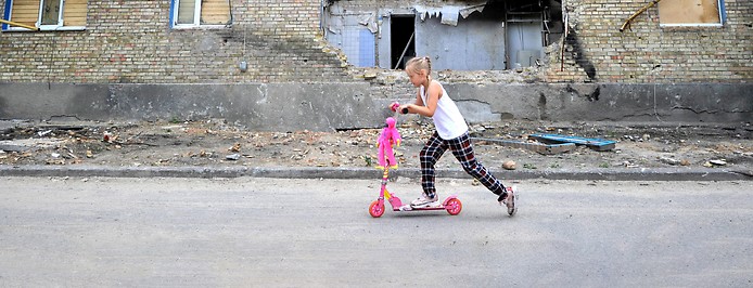 Junges Mädchen auf einem Roller vor einer zerstörten Häuserfront im Dorf Horenka in der Region Kiew